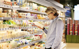 Ein junger Mann steht vor der Kühltheke im Supermarkt. In der Hand hält er eine Milchflasche und liest die Informationen auf dem Etikett durch.