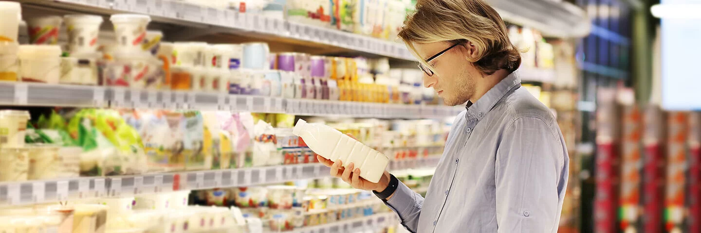 Ein junger Mann steht vor der Kühltheke im Supermarkt. In der Hand hält er eine Milchflasche und liest die Informationen auf dem Etikett durch.