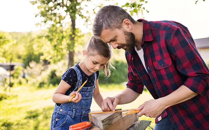 Ein Vater und seine kleine Tochter werkeln im sommerlichen Garten mit Holz. Während der Vater einen Zollstock an das Holzstück anlegt, hält die Tochter einen Bleistift in der Hand.