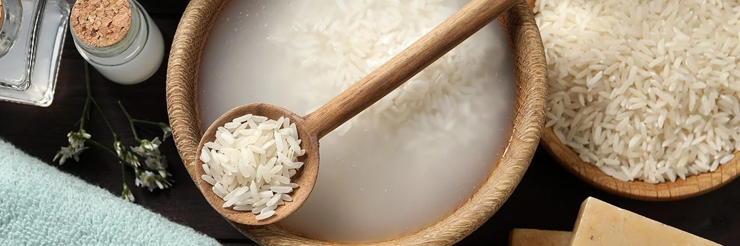 Sicht auf eine Holzschale mit Reis in trübem Reiswasser auf dunklem Grund. Auf der Schale liegt ein mit Reiskörnern gefüllter Holzlöffel.
