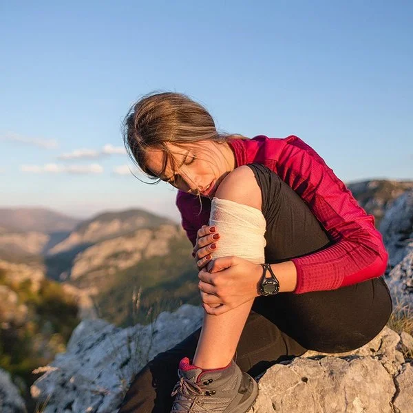 Junge Frau in Wanderkleidung sitzt auf einem Felsen und hält sich den bandagierten Unterschenkel.