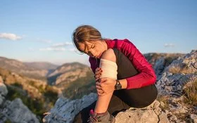 Junge Frau in Wanderkleidung sitzt auf einem Felsen und hält sich den bandagierten Unterschenkel.