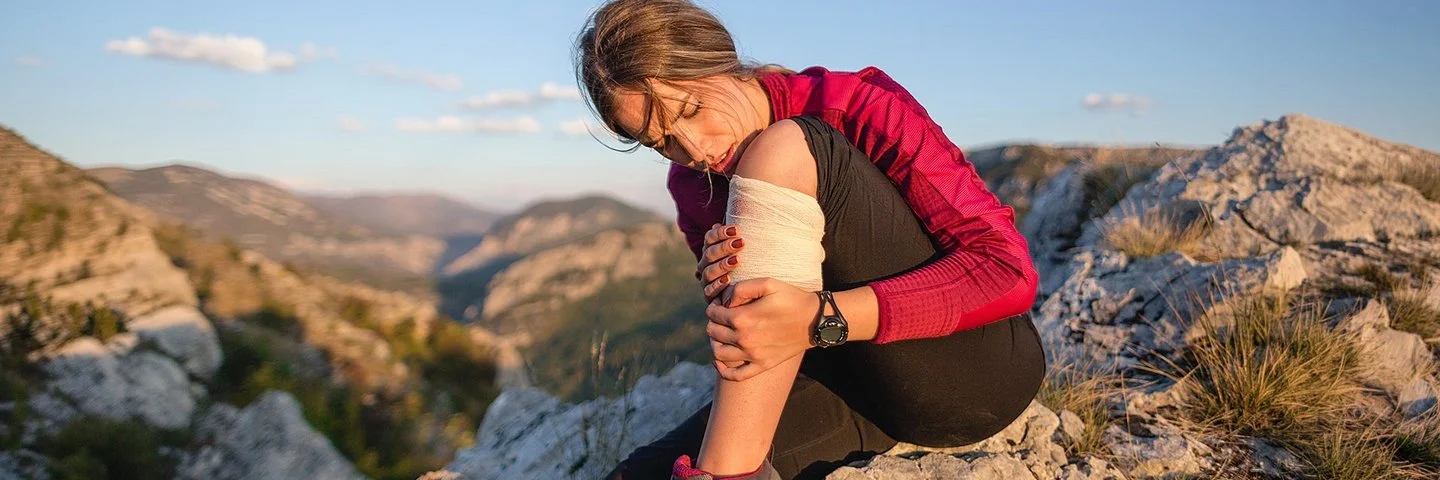 Junge Frau in Wanderkleidung sitzt auf einem Felsen und hält sich den bandagierten Unterschenkel.