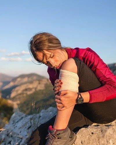 Junge Frau in Wanderkleidung sitzt auf einem Felsen und hält sich den bandagierten Unterschenkel.