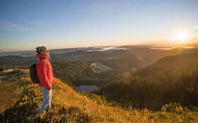 Eine Frau steht auf einem Berg. Vor ihr erstreckt sich der Schwarzwald im Sonnenaufgang. 