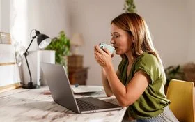 Frau sitzt am Schreibtisch vor einem Laptop und trinkt aus einer Tasse.