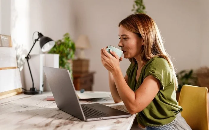 Frau sitzt am Schreibtisch vor einem Laptop und trinkt aus einer Tasse.
