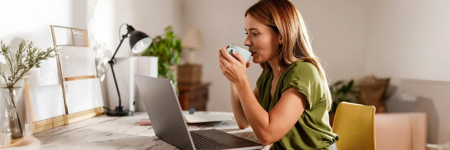 Frau sitzt am Schreibtisch vor einem Laptop und trinkt aus einer Tasse.