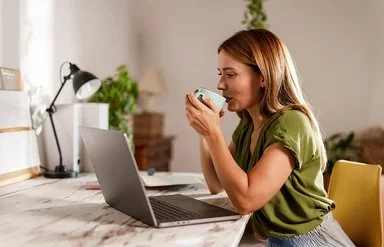Frau sitzt am Schreibtisch vor einem Laptop und trinkt aus einer Tasse.