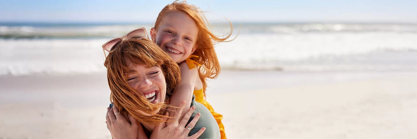 Eine junge Frau trägt ein Mädchen Huckepack an einem Strand, beide lachen fröhlich, der Wind weht ihnen das Haar ins Gesicht.