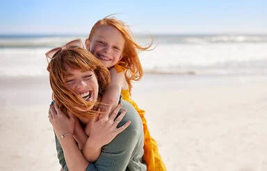 Eine junge Frau trägt ein Mädchen Huckepack an einem Strand, beide lachen fröhlich, der Wind weht ihnen das Haar ins Gesicht.