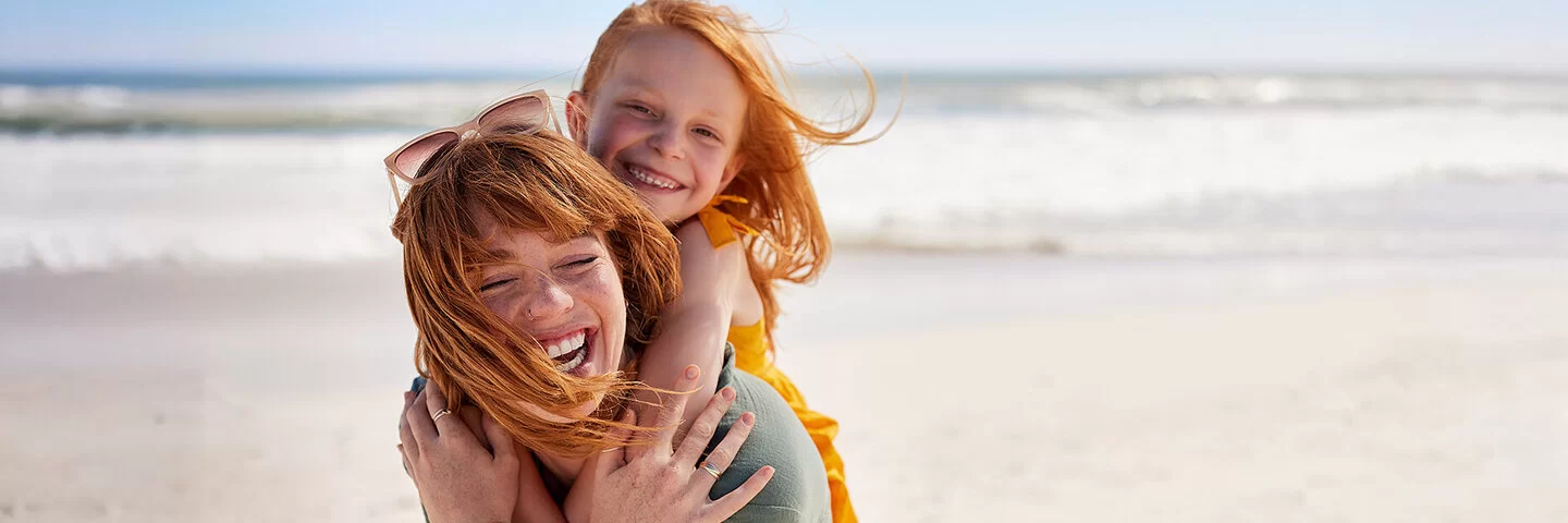 Eine junge Frau trägt ein Mädchen Huckepack an einem Strand, beide lachen fröhlich, der Wind weht ihnen das Haar ins Gesicht.