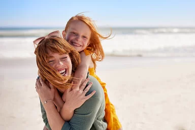 Eine junge Frau trägt ein Mädchen Huckepack an einem Strand, beide lachen fröhlich, der Wind weht ihnen das Haar ins Gesicht.