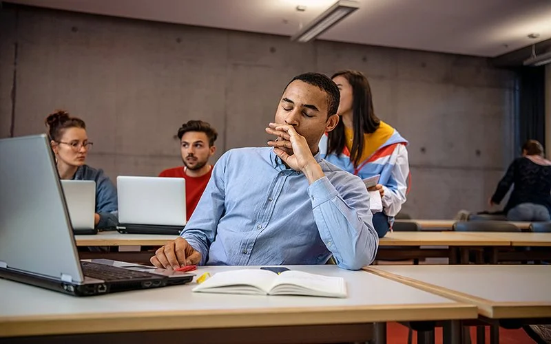 Ein junger Mann sitzt an einem Tisch mit offenem Buch und Laptop und gähnt, im Hintergrund sitzen weitere Personen und arbeiten.
