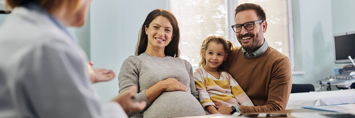 Eine schwangere Frau sitzt neben einem Kind und einem Mann auf Stühlen in einem hellen Raum. Die Frau hält eine Hand auf ihren Bauch. Das Kind sitzt auf dem Schoß des Mannes. Im Vordergrund sitzt eine Person mit weißem Kittel und spricht mit der Familie. Auf dem Tisch liegen Papiere. Im Hintergrund sind ein Wandmonitor und Fenster zu sehen.
