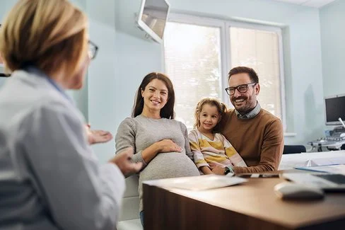 Eine schwangere Frau sitzt neben einem Kind und einem Mann auf Stühlen in einem hellen Raum. Die Frau hält eine Hand auf ihren Bauch. Das Kind sitzt auf dem Schoß des Mannes. Im Vordergrund sitzt eine Person mit weißem Kittel und spricht mit der Familie. Auf dem Tisch liegen Papiere. Im Hintergrund sind ein Wandmonitor und Fenster zu sehen.