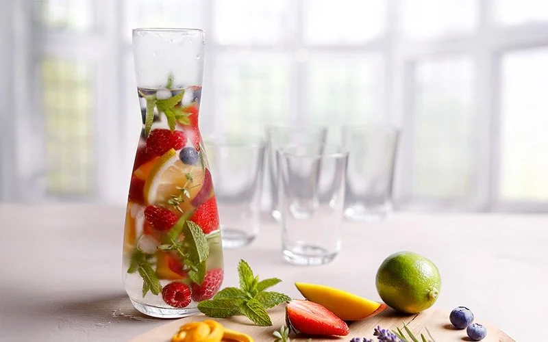 Stilleben einer großen Karaffe selbstgemachten Vitaminwassers mit Früchten, Zitronen und frischen Kräutern darin. Im Vordergrund liegen eine angeschnittene Erdbeere, Mango, Limette, eine Blaubeere, Minze und andere Kräuter. Im Hintergrund sind unscharf vier Gläser zu erkennen.