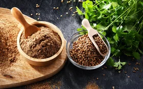 Auf einer schwarzen Tischplatte steht ein Holzbrett, auf dem ein Haufen gemahlener Koriander sowie ein Holzschälchen mit Korianderpulver und einem Schäufelchen liegen. Neben dem Brett befindet sich ein Glasschälchen mit Korianderfrüchten und einem Dosierschäufelchen sowie ein Bund frischer Korianderblätter.
