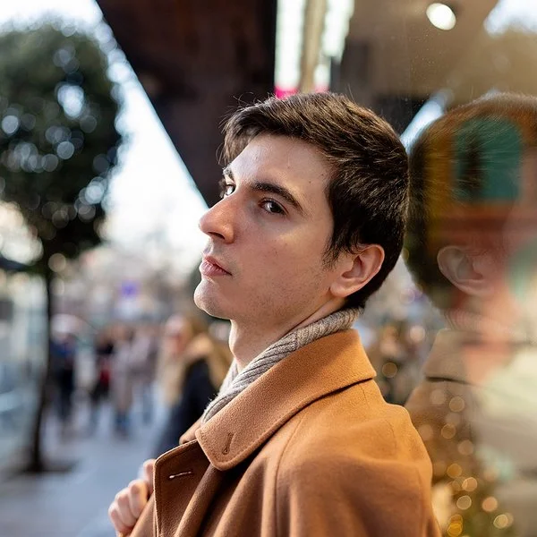 Ein junger Mann steht in einer belebten Stadtstraße und blickt nachdenklich zur Seite. Sein Gesicht spiegelt sich im Schaufenster.