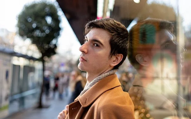 Ein junger Mann steht in einer belebten Stadtstraße und blickt nachdenklich zur Seite. Sein Gesicht spiegelt sich im Schaufenster.