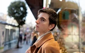 Ein junger Mann steht in einer belebten Stadtstraße und blickt nachdenklich zur Seite. Sein Gesicht spiegelt sich im Schaufenster.