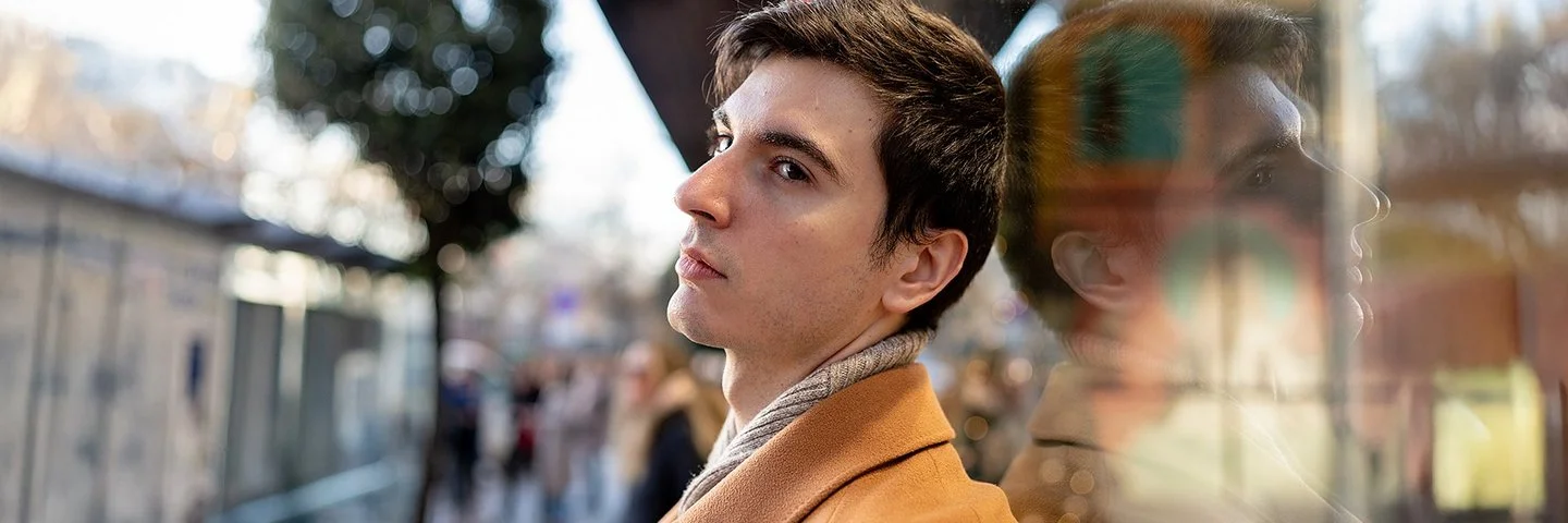 Ein junger Mann steht in einer belebten Stadtstraße und blickt nachdenklich zur Seite. Sein Gesicht spiegelt sich im Schaufenster.