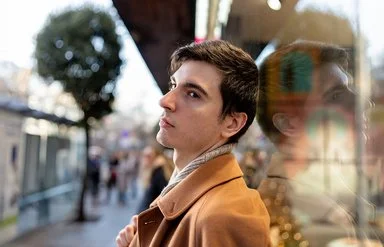 Ein junger Mann steht in einer belebten Stadtstraße und blickt nachdenklich zur Seite. Sein Gesicht spiegelt sich im Schaufenster.