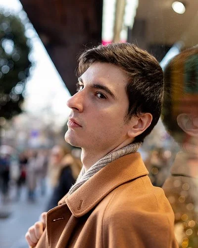 Ein junger Mann steht in einer belebten Stadtstraße und blickt nachdenklich zur Seite. Sein Gesicht spiegelt sich im Schaufenster.