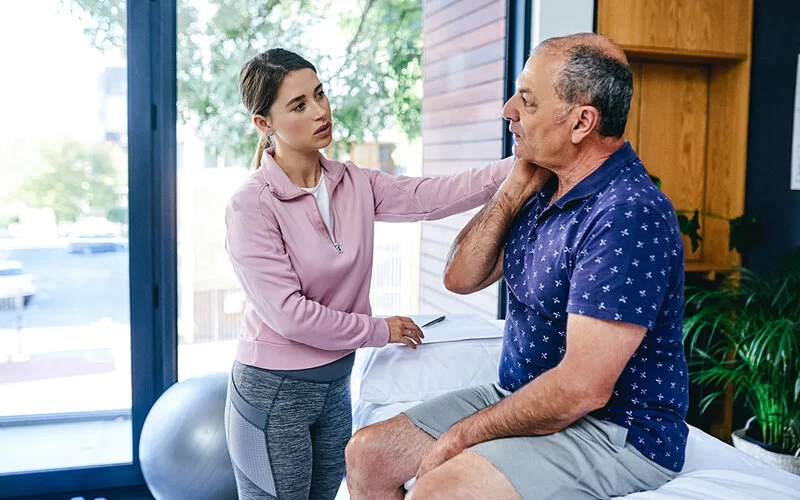 Ein älterer Mann sitzt auf einer Untersuchungsliege und fasst sich mit der rechten Hand an die rechte Halsseite, während eine Physiotherapeutin die besagte Stelle abtastet.