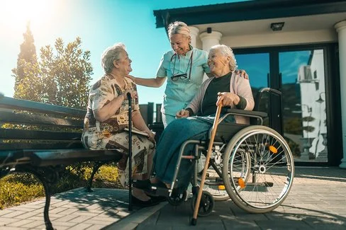 Zwei alte Frauen sitzen draußen in der Sonne. Eine Frau sitzt auf einer Parkbank, die andere im Rollstuhl. Neben ihnen steht eine Pflegerin, die sich mit ihnen unterhält. Im Hintergrund ist der Eingang eines Pflegeheims zu sehen.