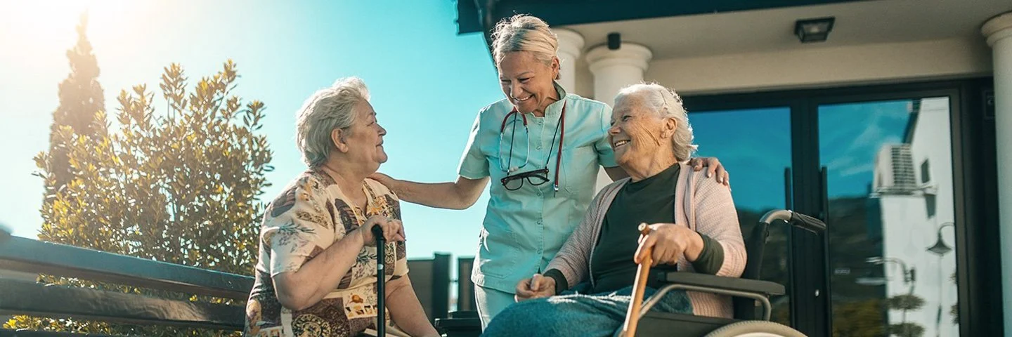 Zwei alte Frauen sitzen draußen in der Sonne. Eine Frau sitzt auf einer Parkbank, die andere im Rollstuhl. Neben ihnen steht eine Pflegerin, die sich mit ihnen unterhält. Im Hintergrund ist der Eingang eines Pflegeheims zu sehen.