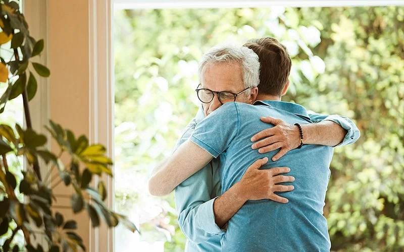 Zwei Männer umarmen sich vor einem Fenster, im Hintergrund sind Bäume zu sehen.