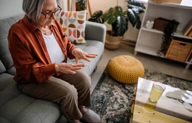 Ältere Dame sitzt auf der Couch im Wohnzimmer, erblickt besorgt ihre Hände.