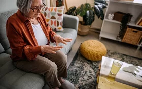 Ältere Dame sitzt auf der Couch im Wohnzimmer, erblickt besorgt ihre Hände.