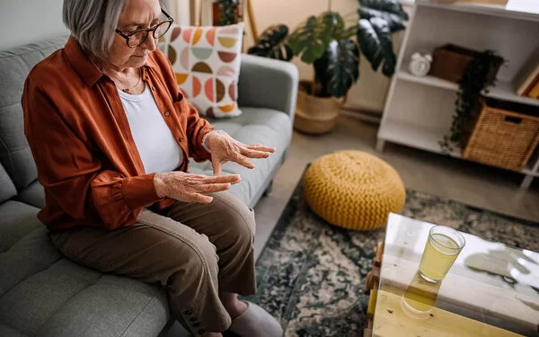 Ältere Dame sitzt auf der Couch im Wohnzimmer, erblickt besorgt ihre Hände.