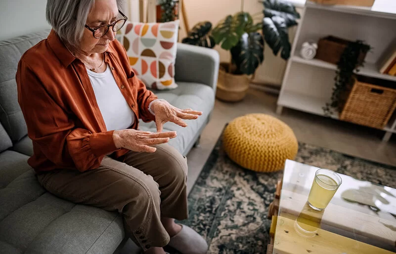 Ältere Dame sitzt auf der Couch im Wohnzimmer, erblickt besorgt ihre Hände.