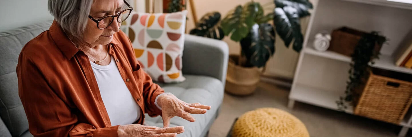 Ältere Dame sitzt auf der Couch im Wohnzimmer, erblickt besorgt ihre Hände.