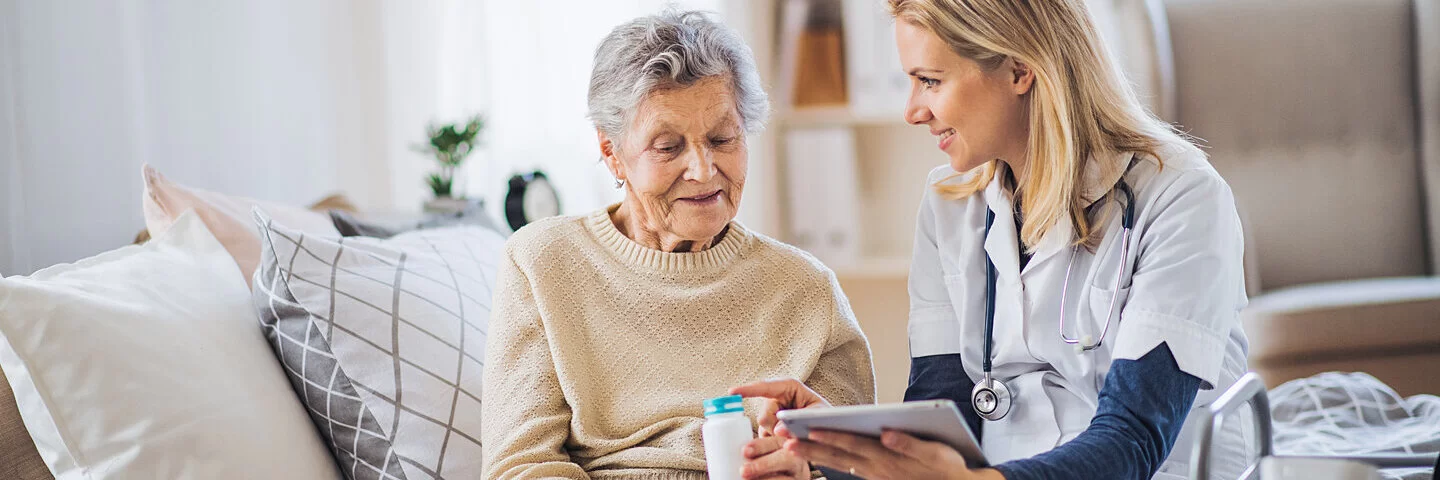 Eine ältere Frau sitzt auf einem Sofa und wird von einer Pflegekraft bei der Einnahme von Medikamenten unterstützt.