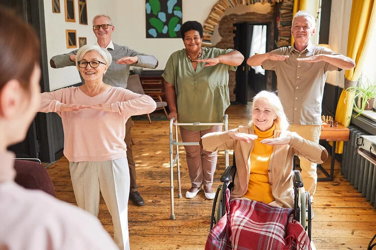 Eine Gruppe von älteren Menschen steht einer Pflegefachperson gegenüber, die Frauen und Männer haben die Arme vor die Brust und ahmen eine Bewegungsübung nach.