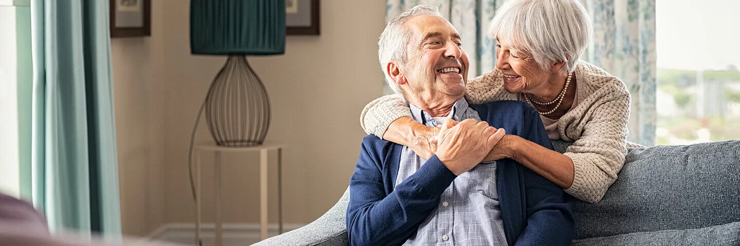 Ältere Frau umarmt ihren Partner über ein Sofa gebeugt.