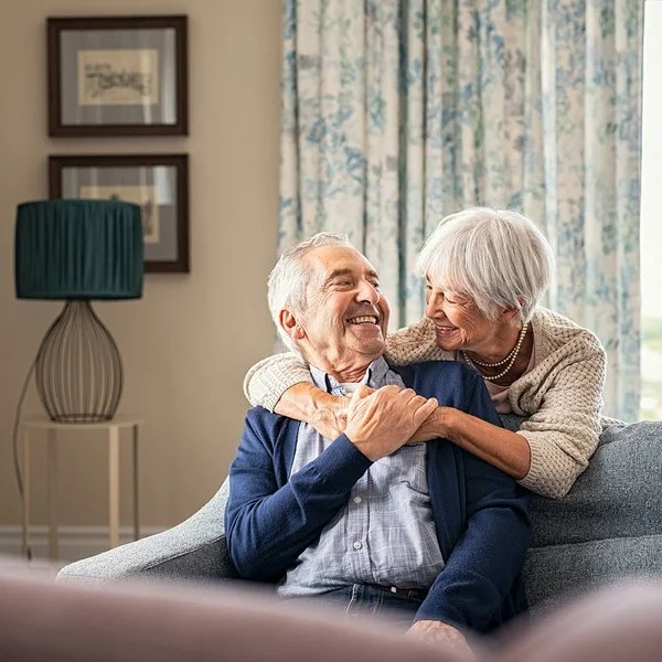 Ältere Frau umarmt ihren Partner über ein Sofa gebeugt.