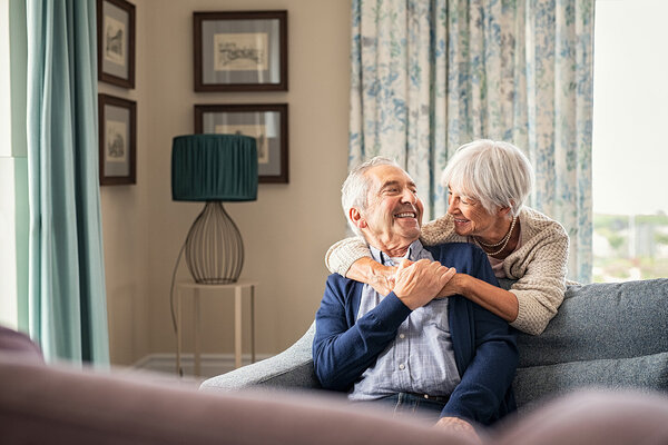 Ältere Frau umarmt ihren Partner über ein Sofa gebeugt.