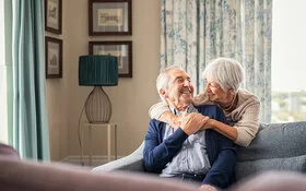 Ältere Frau umarmt ihren Partner über ein Sofa gebeugt.