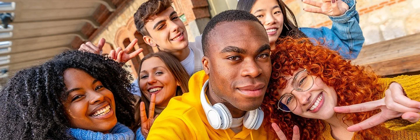 Mehrere junge Erwachsene stehen eng beieinander und posieren für ein Selfie. Eine Person trägt einen gelben Kapuzenpullover und weiße Kopfhörer um den Hals. Im Hintergrund sind ein gemauerter Torbogen und eine Steinwand zu sehen. Einige Personen zeigen das Victory-Zeichen mit den Fingern. Die Szene wirkt lebendig und freundschaftlich.
