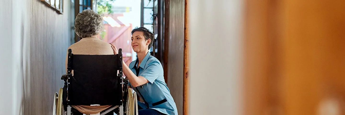 Eine ältere Frau sitzt in einem Rollstuhl und blickt in Richtung der geöffneten Haustür. Neben ihr kniet eine Pflegerin. Sie spricht mit der Frau.