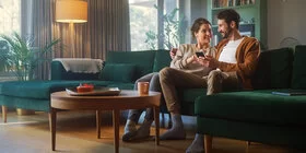 Ein Pärchen in den Dreißigern sitzen nah beieinander auf dem Sofa und lächeln sich an. Der Mann hält ein Smartphone in seiner linken Hand, die Frau zeigt mit ihrer rechten Hand auf das Display.