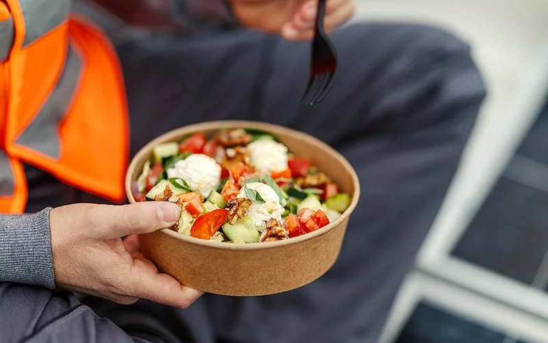 Ein Arbeiter in Sicherheitsweste sitzt neben einem Solarpanel und isst aus einer Pappschale einen gesunden Salat.