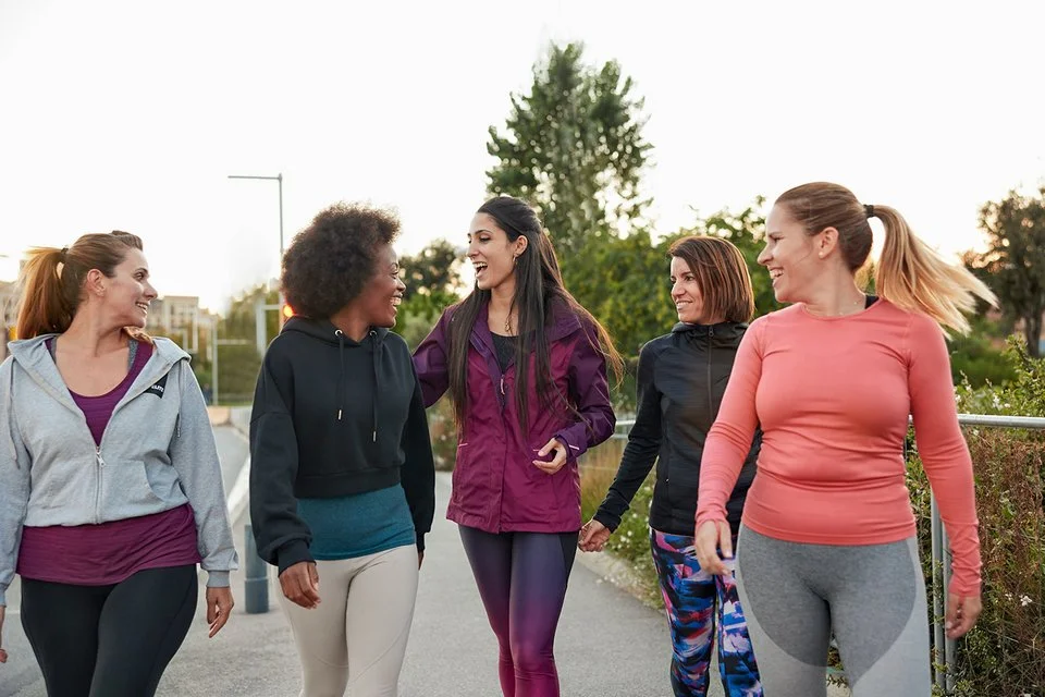 Fünf Frauen sind in Sportkleidung gekleidet. Sie gehen zu einem Sportplatz und unterhalten sich dabei.