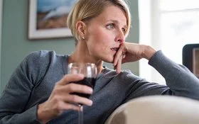 Ein Frau sitzt auf Sofa, nachdenklich den Kopf auf die Hand gestützt und hält ein Glas Rotwein in der anderen Hand
