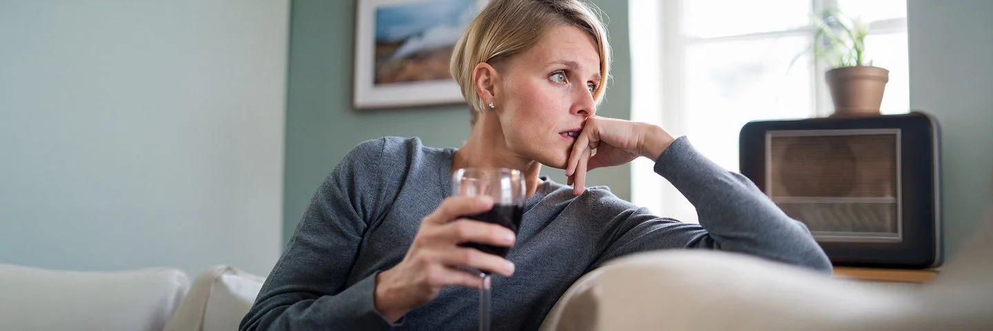 Ein Frau sitzt auf Sofa, nachdenklich den Kopf auf die Hand gestützt und hält ein Glas Rotwein in der anderen Hand